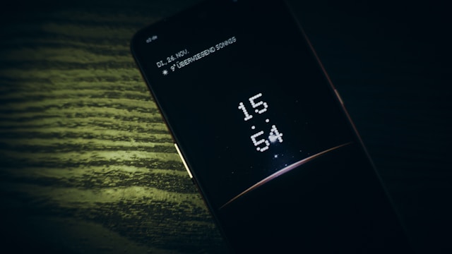 Pametni telefon leži na drvenom stolu i pokazuje digitalno vreme 15:54